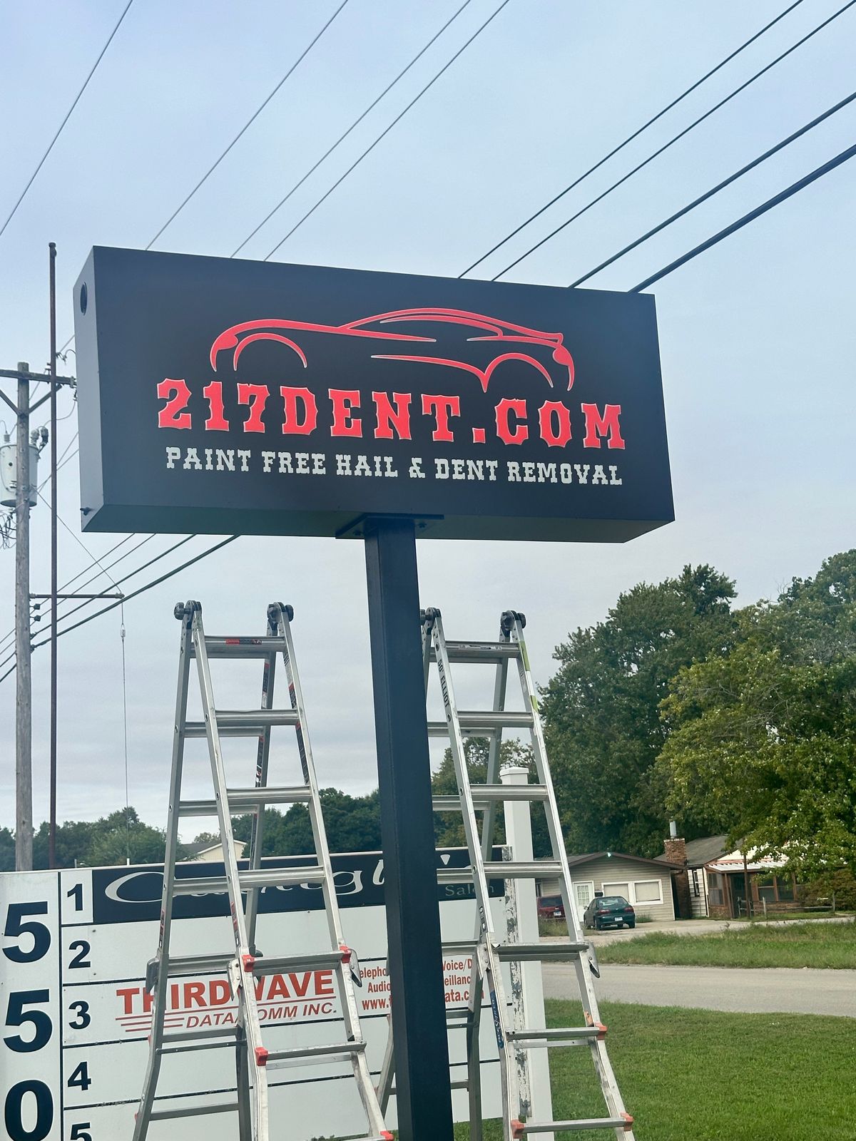 Close-up of the new black 217 Dent pylon sign on a black post, with a red car-outline logo, red 217DENT.COM lettering and white Paint Free Hail and Dent Removal lettering, two aluminum ladders at the base and the multi-tenant 550 address panel beside it