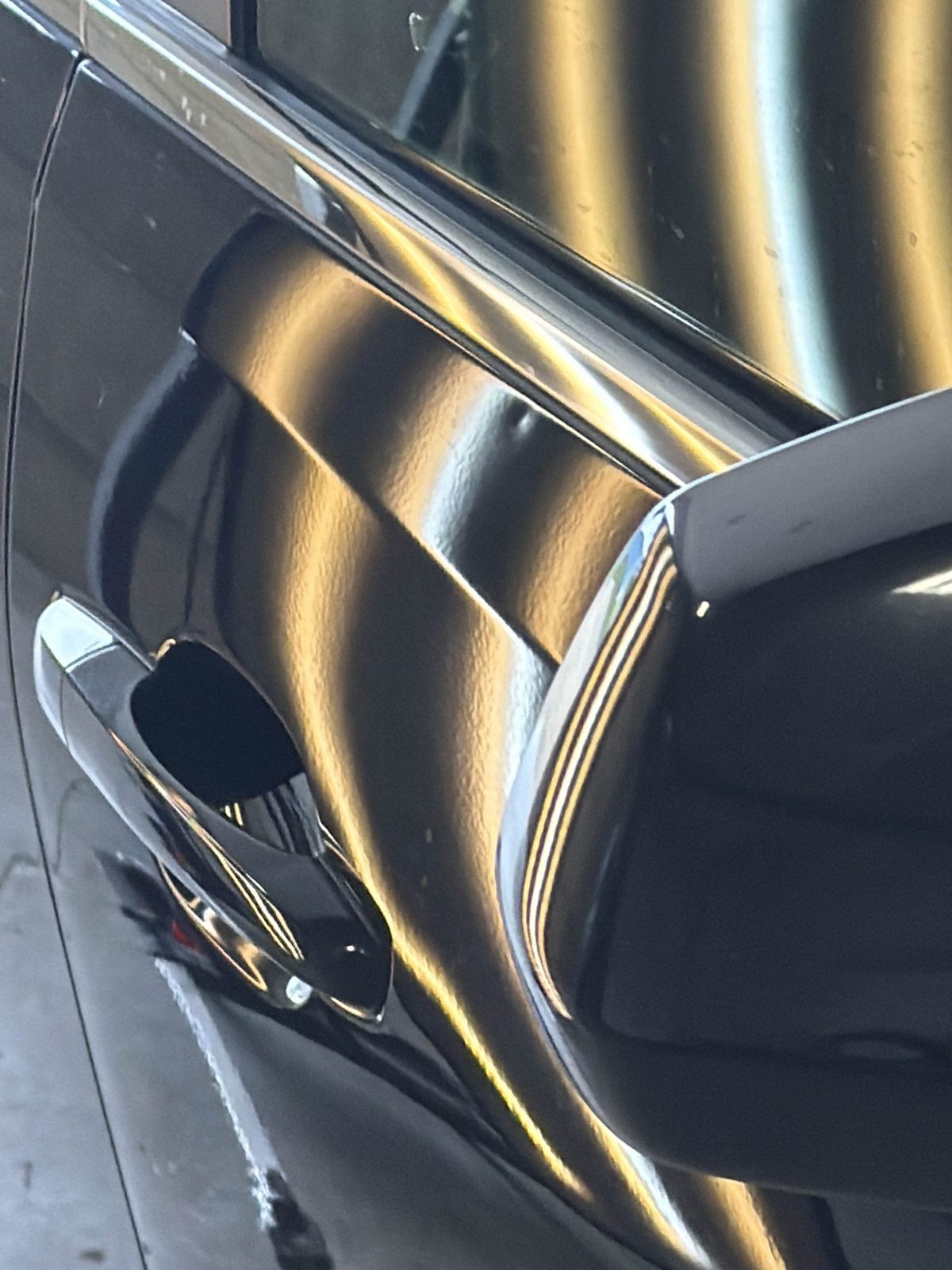 Steel passenger front door of a black Cadillac CTS Sport Wagon near the mirror, with a distorted reflection from hail dents